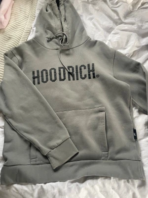 Grå hoodie från Hoodrich - Snygg grå hoodie från Hoodrich med stor svart logga på bröstet. Hoodien har huva med dragsko, magficka och ribbade muddar vid ärmar och nederkant. Perfekt för en chill och streetwear-inspirerad look.