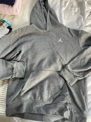 Grå Jordan hoodie med huva - Säljer en grå hoodie från Jordan med klassisk huva och stor magficka. Tröjan har det ikoniska Jumpman/Jordan-loggan broderad i vitt på bröstet. Perfekt för dig som gillar sportig och avslappnad stil. Mjuk och skön bomullsblandning.