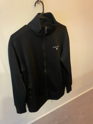 Svart ziptröja från SikSilk - Snygg svart ziptröja från SikSilk med hög krage och diskret logga på bröstet. Tröjan har hel dragkedja framtill och ribbade muddar vid ärmslut och nederkant. Perfekt för en sportig och avslappnad stil.