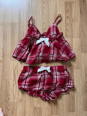 Rödrutigt mjukisset med rosett - Säljer ett par rödrutiga mjukisshorts med vit rosett framtill. Shortsen har elastisk midja och volangkant nedtill. Matchande linne med smala axelband och samma rutiga mönster samt vit rosett framtill ingår.