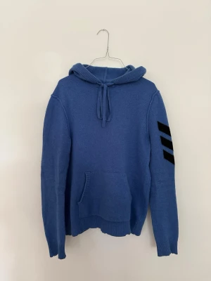 Zadig & Voltaire hoodie i 100 % kashmir, strl M - Snygg blå hoodie med huva och dragsko samt känguruficka framtill. På vänster ärm finns tre svarta ränder som ger en sportig vibe. Tillverkad i mjukt stickat material som känns skönt mot huden. Perfekt för dig som gillar en avslappnad och trendig stil. Pris: 2 400 kr eller bud.