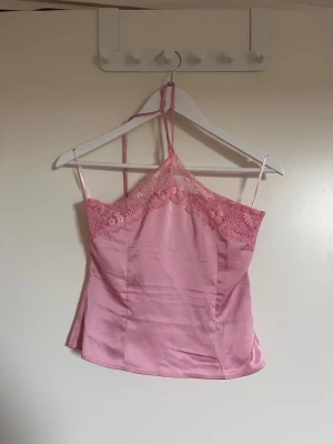 Rosa linnetopp med spetsdetaljer - Söt rosa linnetopp med tunna axelband och spetsdetaljer upptill. Toppen har en halterneck-modell och är gjord i ett glansigt material som ger en lyxig känsla. Perfekt att styla med jeans eller kjol för en trendig look.