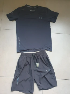Under Armour träningsset svart - Snyggt träningsset från Under Armour i svart, består av en t-shirt och shorts. Tillverkad i lätt och snabbtorkande material med logga på både tröja och byxa. Perfekt för löpning eller gym. Hör gärna av dig om du har frågor eller vill ha fler bilder