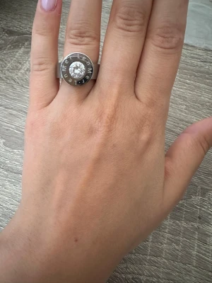 Silverfärgad ring med stor sten från LenMer - Snygg och chunky ring i silverfärgad metall med en stor, rund klar sten i mitten. Ringen har en bred ringband och text ingraverad runt stenen för en modern och cool look. Perfekt statement piece för dig som gillar att sticka ut. Ringen är i storlek 6, alltså 16,5cm i omkrets och aldrig använd med prislapp kvar!