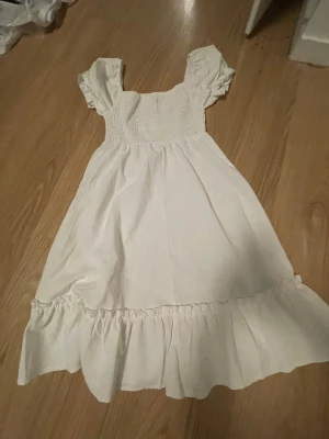 Vit sommarklänning med volang - Söt vit klänning med smock upptill, korta puffärmar och volang längst ner. Klänningen har en romantisk känsla och är perfekt för varma dagar. Tillverkad i ett lätt tyg som känns luftigt och bekvämt. Jättebra till skolavslutning, konfa, eller andra fästliga aktiviteter.