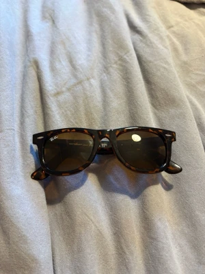 Ray-Ban Wayfarer solglasögon brun - Säljer ett par klassiska Ray-Ban Wayfarer solglasögon med brunmelerat mönster. Snygg retroform med breda bågar och mörka glas. Perfekta för att höja din stil på sommaren eller våren.