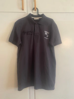 Peak Performance mörkgrå pikétröja,  - Snygg mörkgrå pikétröja med krage och korta ärmar. Tröjan har broderade detaljer på bröstet och klassisk pikéknäppning. Perfekt till jeans eller shorts för en avslappnad look.