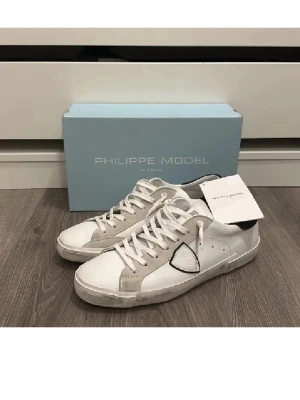 Vita Philippe Model sneakers - Snygga vita sneakers från Philippe Model med grå och svarta detaljer. Skorna har en klassisk låg silhuett, vita snören och en ikonisk sköldlogga på sidan. Tillverkade i läder och mocka för en stilren och trendig look.