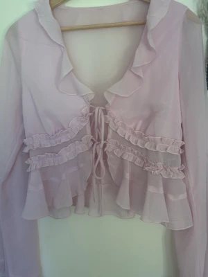 Ljuslila volangblus med knytband - Söt och romantisk blus i ljusrosa transparent tyg med volangdetaljer framtill och längs kragen. Blusen har långa ärmar och knytband framtill. Perfekt för dig som gillar en feminin och trendig stil.
