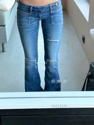 Blå bootcut jeans med slitningar - Säljer ett par blå bootcut jeans med slitningar på knäna och råa detaljer. Jeansen har låg midja, dubbla knappar i linningen och stora framfickor. Perfekta för en avslappnad och trendig look.