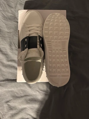 Vita sneakers med mörkblå detalj - Snygga vita sneakers  med bred mörkblå detalj över snörningen. Klassisk låg modell med rund tå och vita sulor. Perfekta för dig som vill ha en stilren och trendig look. Sko lådan följer med den är lite trasig men inget fel på skorna. Strl är 43. En påse följes med