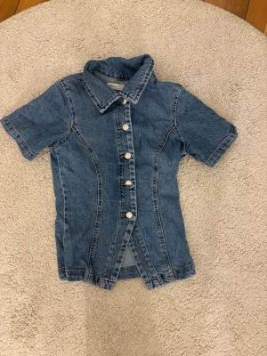 Kortärmad jeansskjorta i denim - Säljer en snygg kortärmad skjorta i klassisk blå denim med knappar framtill och markerad krage. Skjortan har en figurnära passform och coola sömdetaljer som ger en retrovibe. Perfekt att styla till jeans eller kjol för en avslappnad look.