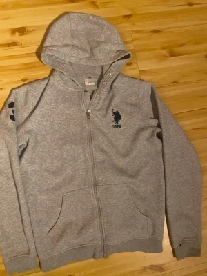 Grå hoodie från U.S. Polo Assn. - Snygg grå hoodie med dragkedja från U.S. Polo Assn. Tryckt logga på bröstet och detaljer på ärmen. Mjuk bomullsblandning, två fickor fram och justerbar huva med metallöljetter. Perfekt för en avslappnad och sportig stil. Använd 2-3 gånger