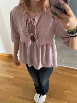 Rosa blus med knyt och trekvartsärm - Söt rosa blus med två knyt framtill och lätt rynkad midja. Blusen har trekvartsärmar och ett luftigt, mjukt tyg i bomull. Perfekt för dig som gillar romantisk och avslappnad stil.