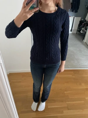 Mörkblå kabelstickad tröja - Snygg mörkblå kabelstickad tröja med rund halsringning och långa ärmar. Tröjan har ett klassiskt flätmönster och ribbade muddar vid ärmslut och nederkant. Perfekt att matcha med jeans för en avslappnad look. Tyget kan kännas lite sticksigt!
