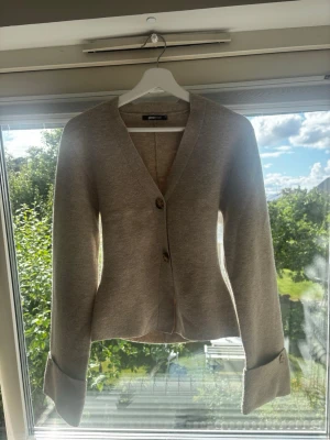 Beige kofta från gina  - Stilren beige kofta med två knappar framtill och breda muddar vid ärmsluten. Koftan har v-ringning och rak passform, perfekt att slänga över en topp. Tillverkad i mjukt stickat material som känns skönt mot huden.