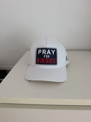 Vit keps med 'PRAY FOR BIRDIES'-patch - Snygg vit keps med rak skärm och stor patch framtill där det står 'PRAY FOR BIRDIES' i vitt och rött på mörkblå bakgrund. Sidan har ett diskret svart logomärke och materialet är lätt och ventilerande med små hål på sidorna. Använd 2ggr
