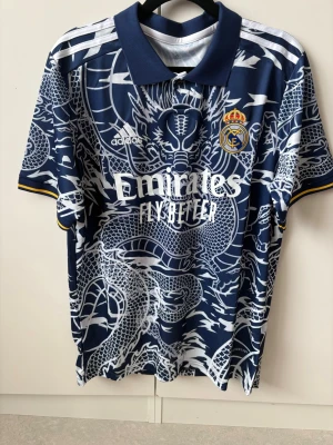 Real Madrid blå draktröja Adidas - Säljer en blå Real Madrid matchtröja med unikt drakmönster i vitt. Tröjan har krage, korta ärmar och gula detaljer vid ärmsluten. Officiell klubb- och Adidaslogga på bröstet samt Emirates Fly Better-tryck framtill. Perfekt för dig som älskar fotboll och vill sticka ut.