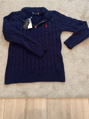 Mörkblå kabelstickad tröja Polo Ralph Lauren - Snygg mörkblå kabelstickad tröja från Polo Ralph Lauren med halv dragkedja och röd broderad logga på bröstet. Tröjan har ribbade muddar och hög krage, perfekt för lager på lager. Klassisk och tidlös design som passar till det mesta.