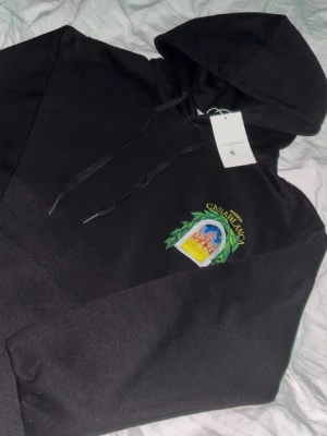  Casablanca hoodie - Svart hoodie med stor färgglad tryck på ryggen och mindre tryck på bröstet. Motivet visar en byggnad genom ett fönster med gröna blad runt och texten 'Avenida Casablanca'. Huva med dragsko och känguruficka framtill. Perfekt för dig som gillar unika prints.