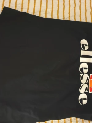 Svart baddräkt från Ellesse - Snygg svart baddräkt från Ellesse med stort vitt och orange logotryck framtill. Klassisk modell med sportig känsla, perfekt för stranden eller poolen. Materialet är stretchigt och följsamt för bästa komfort.