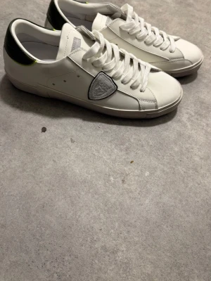 Vita sneakers med gröna detaljer - Snygga vita sneakers från Philippe Model med gröna detaljer på hälen och en unik patch på sidan. Skorna har vita snören och är tillverkade i slätt läder för en clean look. Perfekta för dig som gillar stilrena men ändå unika sneakers.