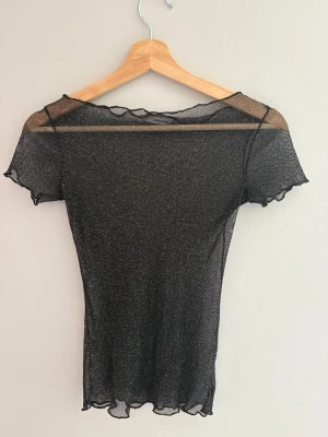Svart glittrig mesh topp - Snygg svart glittrig mesh topp med korta ärmar och vågiga kanter vid ärmslut och nederkant. Tunt och transparent material som ger en cool och trendig look. Aldrig använd endast testad. Skriv privat för mer information. 
