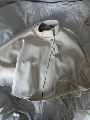 Ljusbeige kappa med bälte - Stilren ljusbeige kappa med ståkrage och knäppning framtill. Kappan har ett matchande bälte i midjan och raka ärmar. Materialet ser ut att vara mjukt och något tjockare, perfekt för kyligare dagar. Enkel och modern design som passar till många outfits.