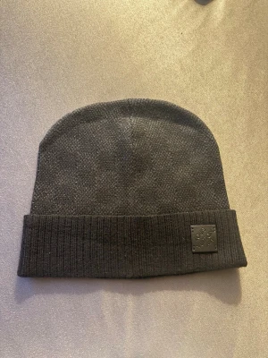 Mörkgrå mössa  - Snygg mörkgrå mössa med uppvikt kant och en svart patch med logotypen framtill. Tillverkad i mjukt stickat material som håller dig varm och stilren under kalla dagar.