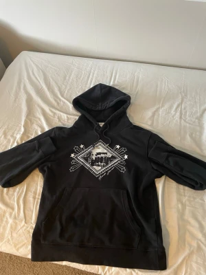Yves Saint Laurent Hoodie - Säljer en svart hoodie från saint Laurent med ett stort vitt grafiskt tryck på bröstet. Hoodien har huva med dragsko och en klassisk magficka. Materialet känns mjukt och bekvämt, perfekt för chill dagar. Passar dig som gillar streetwear och enkel stil.