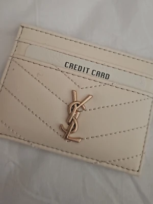 Beige kortfodral  - Snyggt beige kortfodral i läder med guldfärgad  framtill. Kortfodralet har flera fack för kreditkort och stilrena sömmar i chevronmönster. Perfekt för dig som vill ha ett lyxigt och smidigt sätt att bära dina kort.