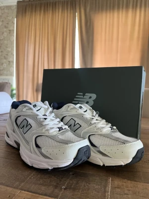 New Balance 530 vita sneakers - Snygga New Balance 530 sneakers i vitt med silver- och marinblå detaljer. Ovandelen är i mesh och syntetmaterial för en sportig look. Klassisk N-logga på sidan och ABZORB-dämpning i sulan för extra komfort. Perfekta för en trendig och avslappnad stil.