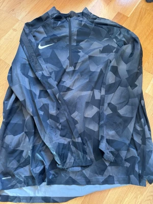 Nike Windrunner med camomönster - Nike Windrunner med svart och grått camomönster, halv dragkedja framtill och lång ärm. Tillverkad i lätt och vindtätt material med Nike Shield-teknologi. Perfekt för löpning eller träning utomhus när det blåser.