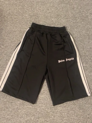 Palm Angels svarta shorts med vita ränder - Palm Angels shorts i svart med vita sidoränder och logga broderad på vänster ben. De har elastisk midja och är tillverkade i ett glansigt, sportigt material som polyester. Perfekta för en avslappnad streetwear-look.
