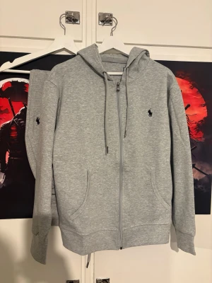 Grå loungewear-set från Polo Ralph Lauren - Mysigt grått loungewear-set från Polo Ralph Lauren med zip-hoodie och matchande byxor. Setet har diskreta svarta loggor på bröstet och benet, dragsko i midjan och huva samt ribbade muddar. Perfekt för chill dagar hemma.