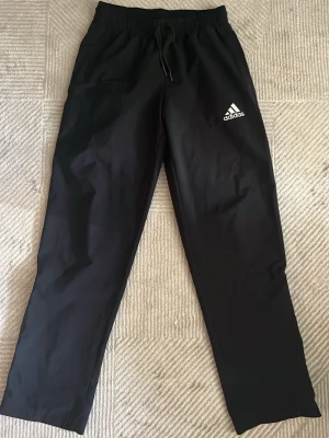 Svarta träningsbyxor från Adidas - Snygga svarta träningsbyxor från Adidas med klassisk logga på vänster lår. Byxorna har rak passform, resår i midjan med dragsko och är tillverkade i ett lätt och bekvämt material som passar perfekt till träning eller chill.