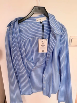 Blåvit randig skjorta från Bershka - Snygg blå skjorta med vita ränder från Bershka. Skjortan har klassisk krage, knäppning framtill och långa ärmar. Perfekt för en trendig och avslappnad look. Tillverkad i ett lätt och bekvämt material.