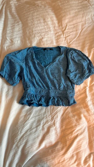 Blå blus med volang och puffärm - Söt blå blus med små vita prickar, v-ringad hals och puffiga korta ärmar. Modellen är croppad med volangkant nedtill och resår i midjan. 
