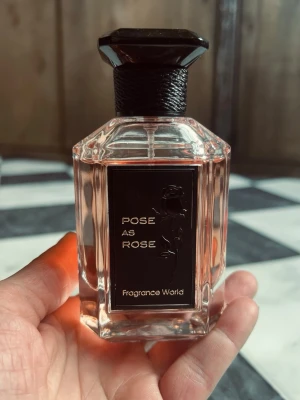 Pose As Rose Fragrance World, 100ml - Pose As Rose från Fragrance World är en parfym i en elegant, sexkantig glasflaska med ljusrosa innehåll. Flaskan har en svart etikett med guldfärgad text och en rosdetalj, samt ett svart, sexkantigt lock med snördetalj runt halsen.