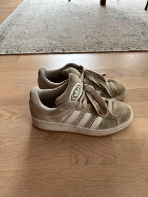 Beige Adidas Campus - Beiga Adidas campus  sneakers i mocka med klassiska vita ränder på sidorna och Adidas-logga på plösen. Skorna har en låg profil och matchande beige snörning. 