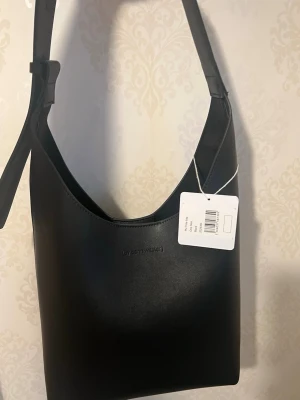 Snygg väska oanvänd ! - Stilren svart axelväska från Carin Wester i slätt konstläder. Väskan har en enkel, rektangulär form med bred axelrem och diskret logga framtill. Perfekt för dig som gillar minimalistisk design och vill ha en väska som rymmer det viktigaste.