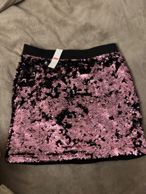 Rosa paljettkjol  - Glittrig kortkjol täckt av rosa paljetter med svart midjeresår. Kjolen är perfekt för fest och den går även att vika ner i midjan