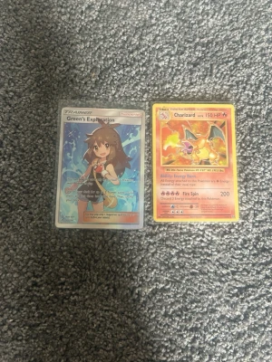 Två Pokémonkort Charizard & Green - Säljer två Pokémonkort: Charizard med röd och gul ram samt Green’s Exploration med holografisk effekt. Båda korten har klassisk Pokémon-baksida och är perfekta för samlare eller spelare. Kortens motiv är färgstarka och detaljerade.