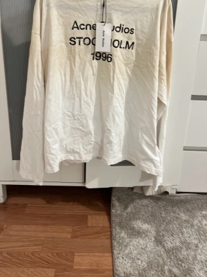 Vit långärmad t-shirt Acne Studios - Vit långärmad t-shirt från Acne Studios med svart tryck framtill där det står 'Acne Studios STOCKHOLM 1996'. Klassisk passform och rund halsringning. Perfekt för en clean och stilren look.
