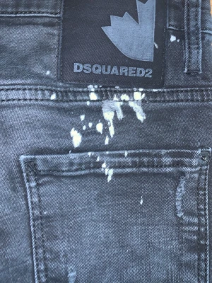 Svarta Dsquared2 jeans med slitningar - Säljer ett par svarta jeans från Dsquared2 med coola slitningar och vita färgstänk. Jeansen har en avslappnad passform, klassiska fickor och en snygg patch bak i midjan. Materialet är denim med stretch för extra komfort.