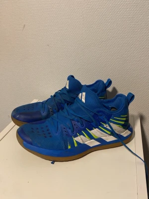 Blåa Adidas inomhusskor - Säljer ett par blåa Adidas inomhusskor med vita och limegröna detaljer samt gummisula. Skorna har ventilerande meshmaterial och snörning för bra passform. Perfekta för sportaktiviteter inomhus.