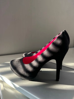 Svarta pumps med rosa innersula - Säljer ett par klassiska svarta pumps med hög klack och rundad tå. Skorna har en snygg rosa innersula som ger en cool kontrast. Materialet ser ut att vara mockaimitation vilket ger en mjuk och matt finish. Perfekta för dig som vill sticka ut lite extra.