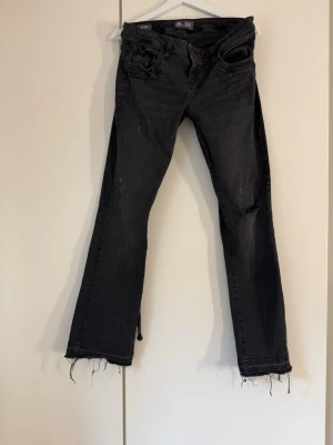 Svarta bootcut jeans med rå kant - Säljer ett par svarta bootcut jeans med slitningar och råa, fransiga benslut. Jeansen har fem fickor och en avslappnad passform. Perfekta för dig som gillar en edgy och trendig stil.