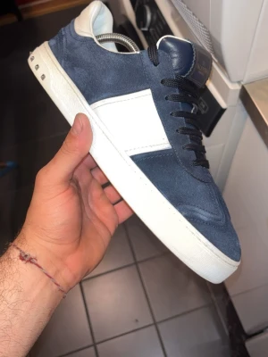 Blå och vit sneaker i mocka - Snygg sneaker i mörkblå mocka med vit läderdetalj på sidan och vit sula. Skon har svarta skosnören och en klassisk låg profil. Perfekt för dig som gillar stilrena och trendiga sneakers med en sportig touch.