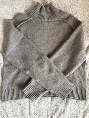 Grå stickad polotröja från H&M - Mysig grå stickad polotröja från H&M i storlek XS. Tröjan har ribbade muddar vid ärmslut och nederkant samt en hög, ribbad polokrage. Perfekt för kalla dagar och enkel att matcha med olika outfits.